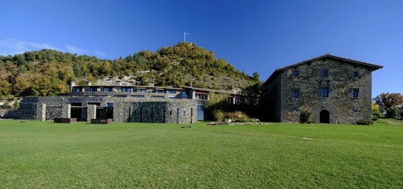 Vista de una montaña con una cruz en la cima, un gran edificio de piedra en el primer plano y un campo de césped verde