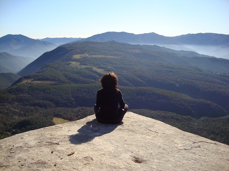 Una persona sentada en una roca en la cima de una montaña, mirando hacia un paisaje de montañas verdes y el cielo azul.