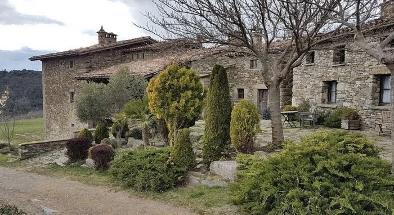 Casa de piedra con ventanas y pequeños objetos de mobiliario en el patio, rodeada de árboles y arbustos en un paisaje rural.