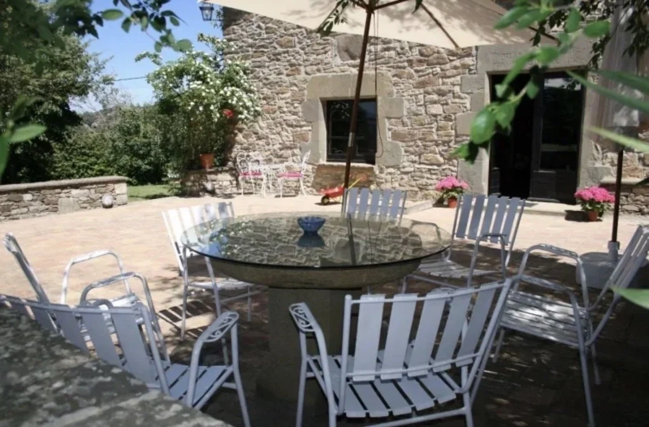Muebles de patio con mesa de cristal y sillas blancas, rodeado de plantas y flores, en un patio con paredes de piedra y sombrillas