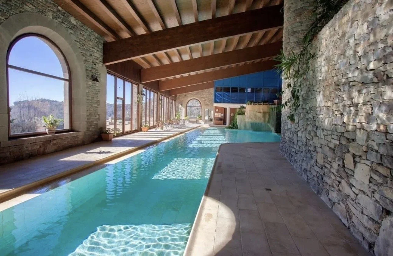Interior de una alberca cubierta con paredes de piedra y ventanas grandes, con techos de madera.