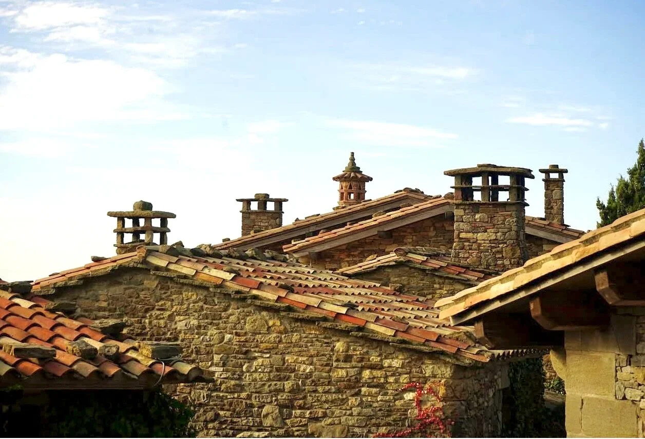 Tejados de casas tradicionales de piedra con chimeneas de barro y tejas de cerámica en un valle, bajo un cielo despejado.
