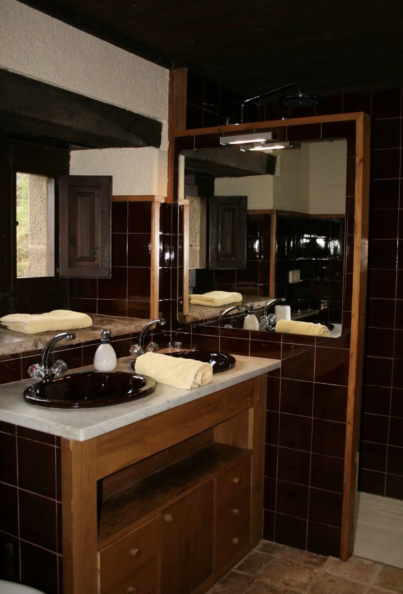 Este es un baño con lavabo doble, espejo grande, paredes de azulejos y gabinetes de madera, toallas enrolladas sobre el lavabo y un reflejo que muestra una ventana.