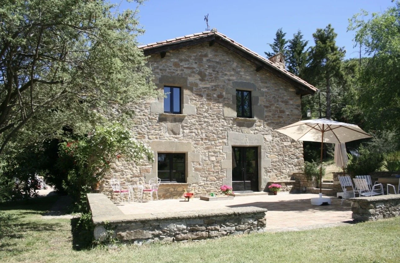 Casa de piedra con ventanas y puerta, terraza con sombrilla y sillones, rodeada de árboles y jardín