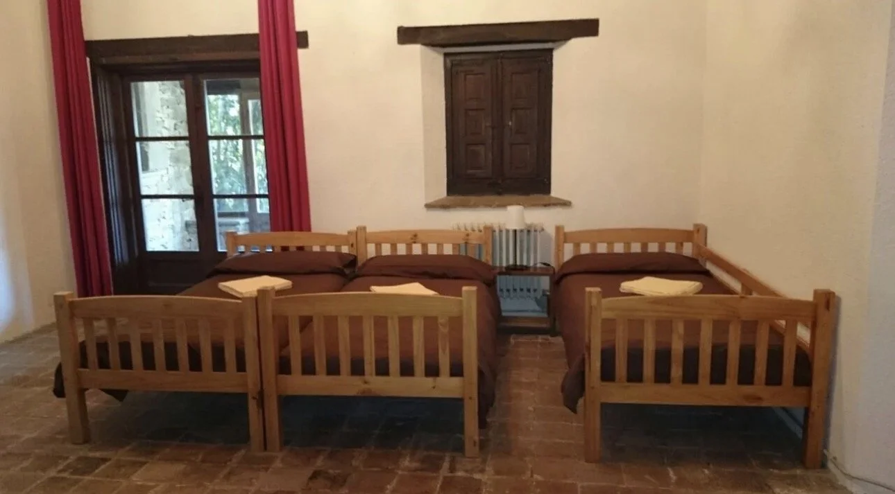 Cuatro camas individuales con ropa de cama marrón en una habitación con paredes blancas, cortinas rojas y ventanas de madera con vista a un patio con paredes de piedra.