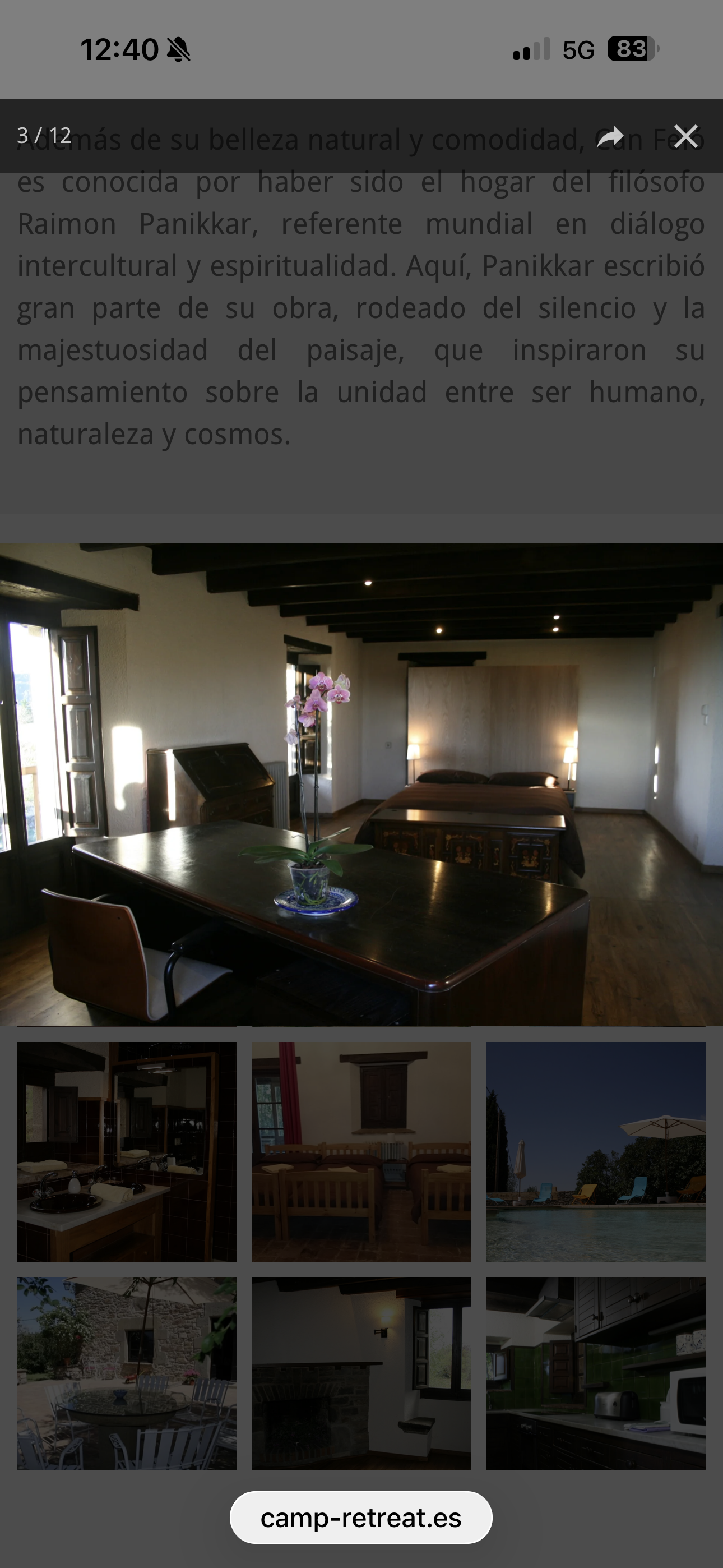 Una habitación de hotel con una cama, mesas y lámparas decorativas. Ventanas con cortinas rojas, piso de madera y paredes blancas. Sol y naturaleza visible afuera.