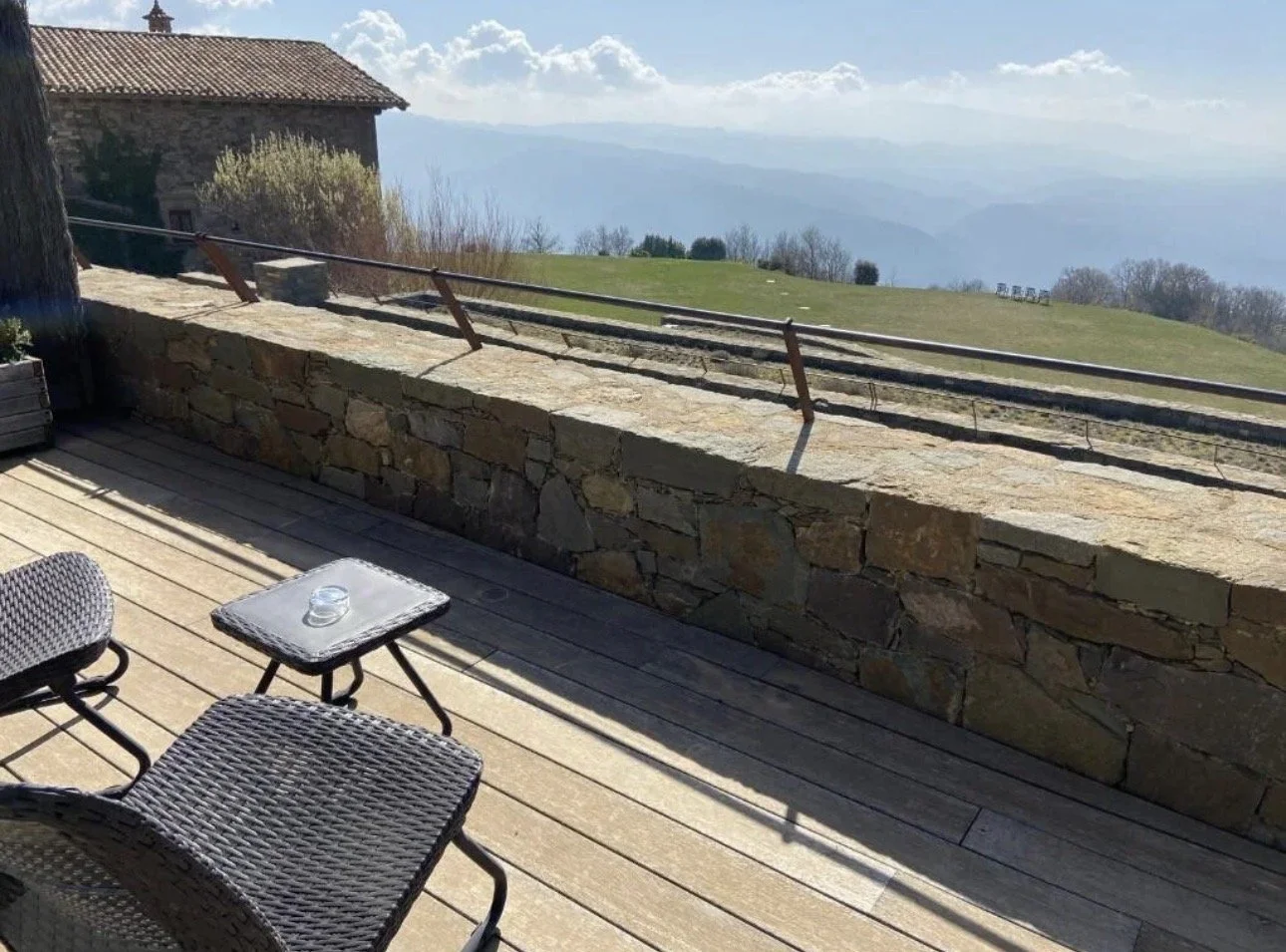Vista de un balcón con muebles de mimbre y una pequeña mesa con un vaso, con un muro de piedra que da hacia un paisaje de montañas y campo bajo un cielo parcialmente nublado.
