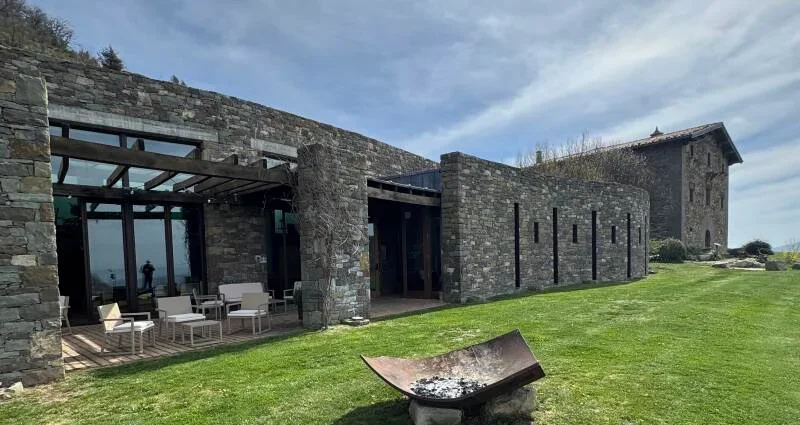 Edificio moderno de piedra con ventanas grandes y patio con muebles, en un espacio con césped y un quemador de fuego, bajo un cielo nublado.