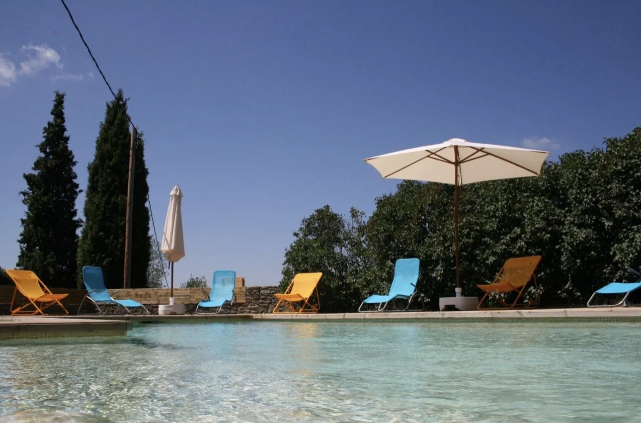 Piscina con sillas de playa y sombrillas, árboles y cielo despejado en el fondo.