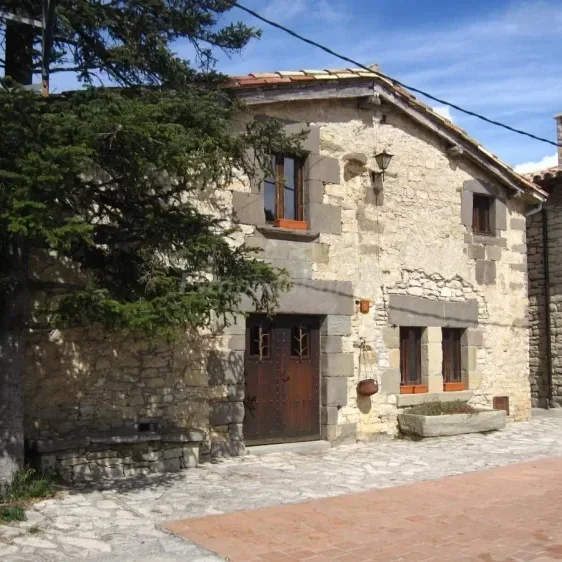 Casa de piedra con ventanas de madera y puertas, en un entorno rural, con un árbol cercano y calles empedradas.