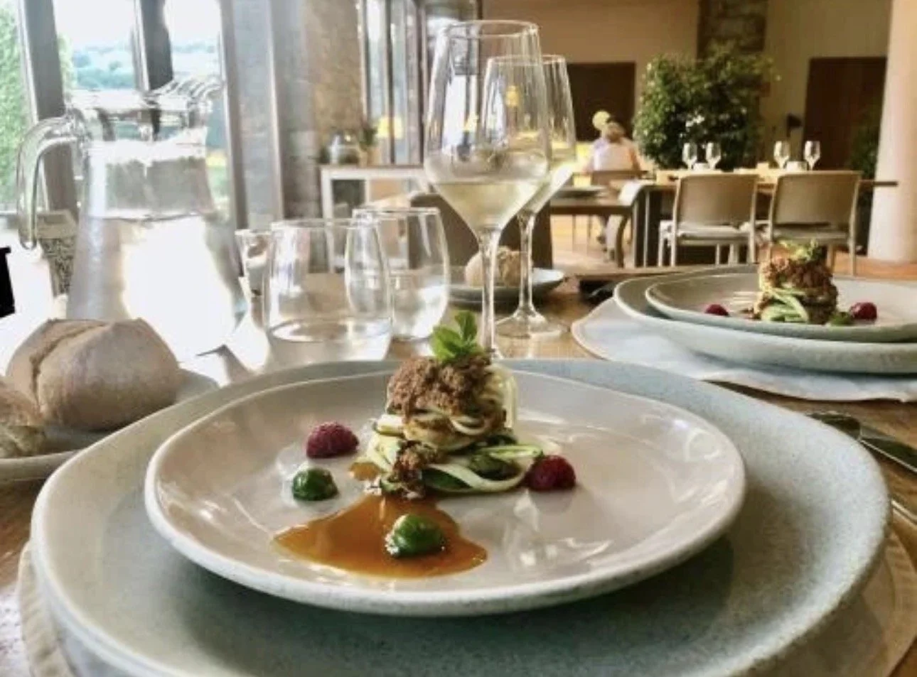 Plato de comida gourmet con carne, verduras y salsa, en un restaurante elegante con copas y agua en la mesa.