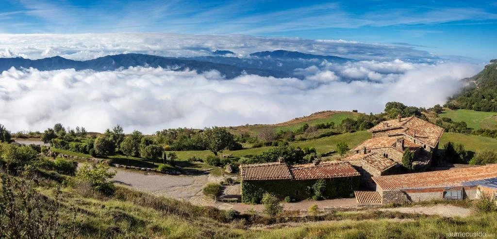 Paisaje de montañas con casas de techos de tejas, vegetación y nubes en el cielo.