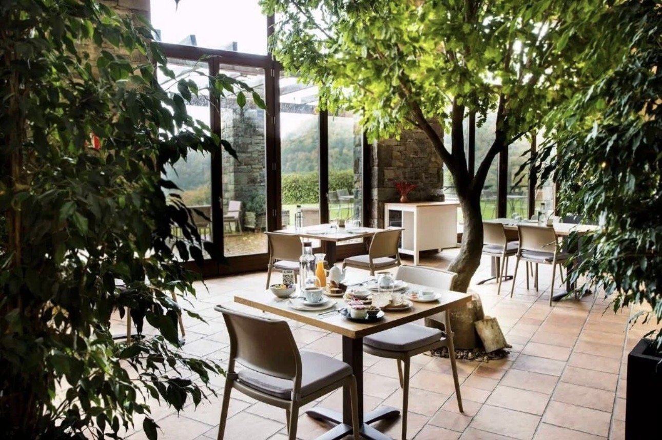 Interior de un restaurante con mesas, sillas y árboles, con vistas al exterior a través de ventanas grandes.