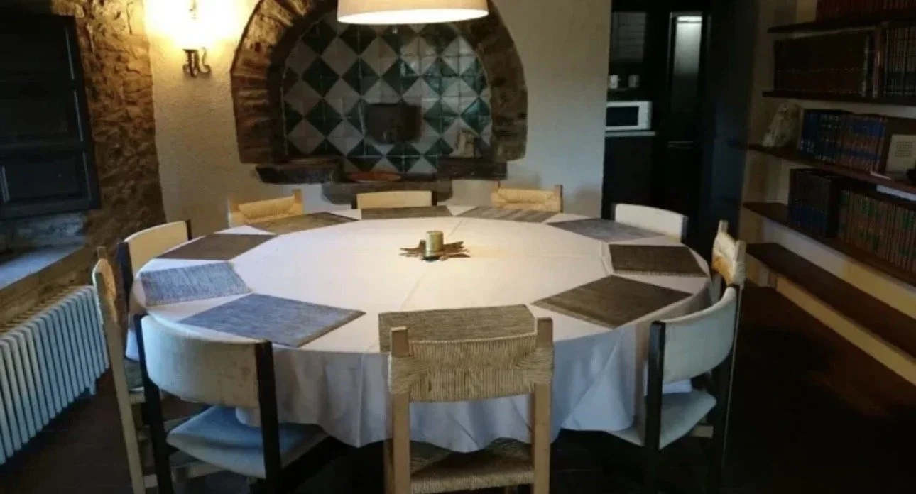 Mesa redonda con mantel blanco y ocho sillas alrededor, en una sala con paredes de piedra y una alcoba decorada en tonos oscuros, con libros en estantes a la derecha y una chimenea o revestimiento de ladrillo en el fondo.