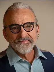 Close-up of an older man with gray hair and beard wearing glasses, a blue shirt, and a dark sweater, smiling slightly.