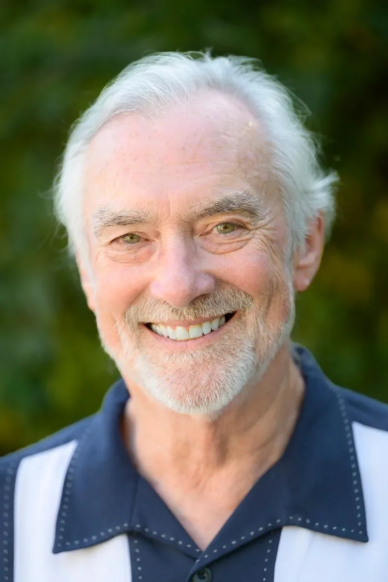 Close-up portrait of a smiling elderly man with white hair and beard, wearing a dark collared shirt with white accents, standing outdoors with blurred green foliage in the background.