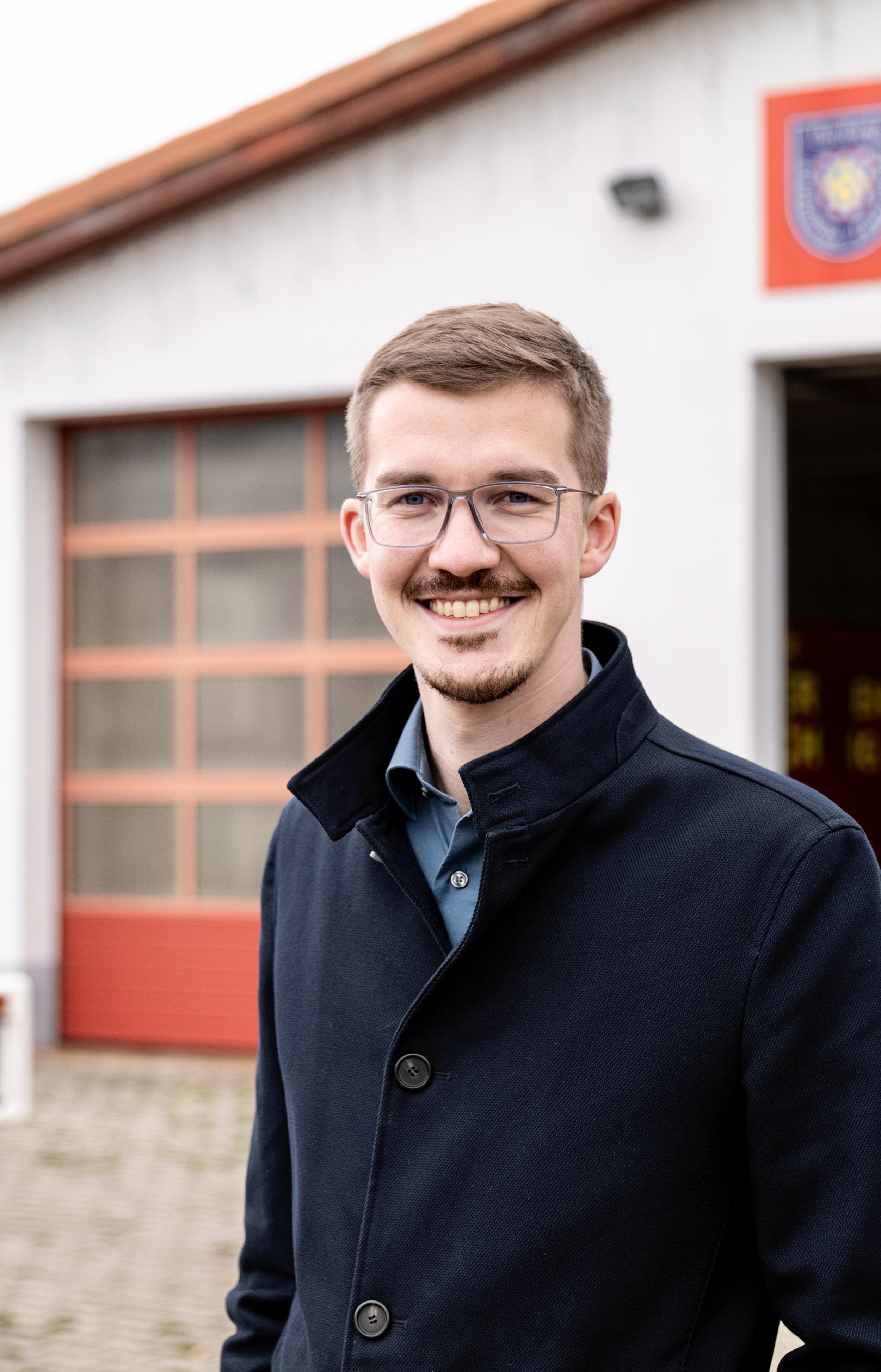 Junger Mann mit Brille, dunkler Mantel und blauem Hemd vor einer Feuerwache mit rotem Tor im Hintergrund.