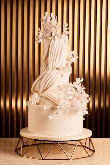 Elegant white tiered wedding cake with white floral decorations on a round stand against a gold vertical striped background.
