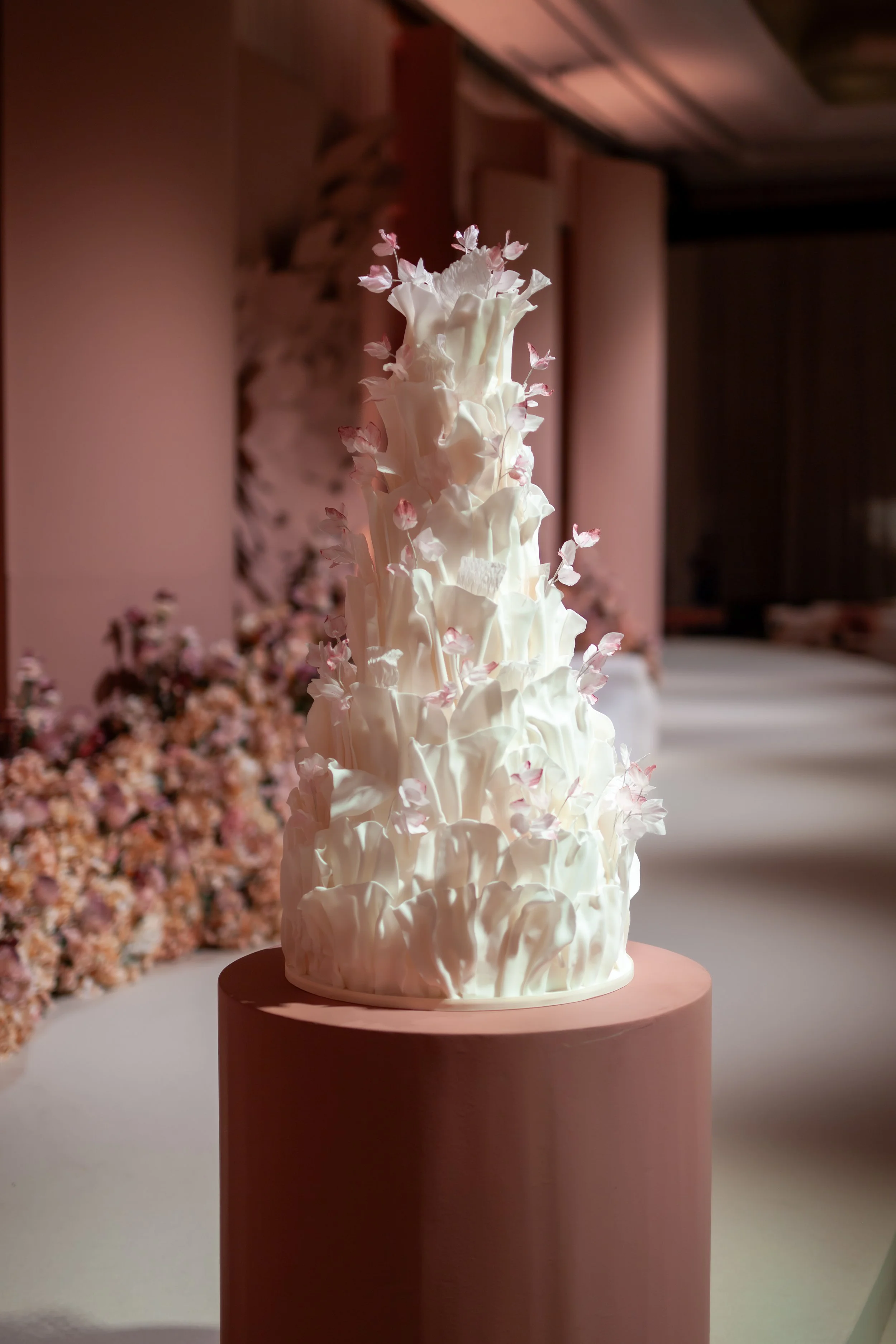 Tall multi-tier white wedding cake with cascading pink and white sugar flowers, displayed on a soft pink pedestal.