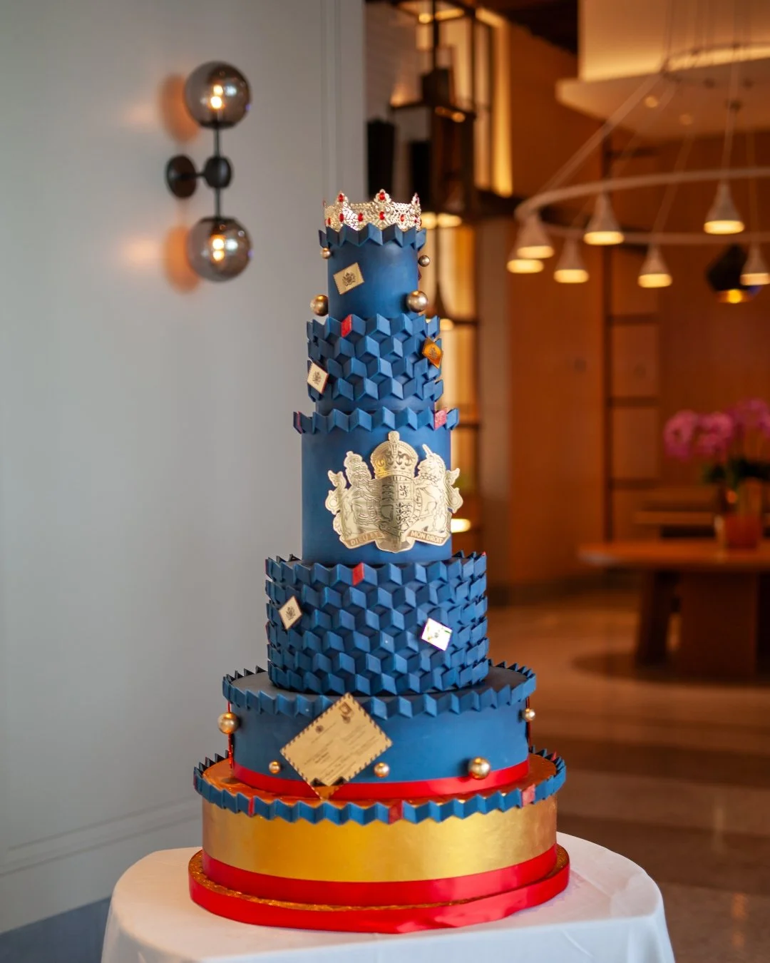 Tall multi-tier celebration cake with bold blue geometric detailing,  Royal Crest, gold accents and a crown topper for HRH King Charles 3rd Birthday and The British Embassy.