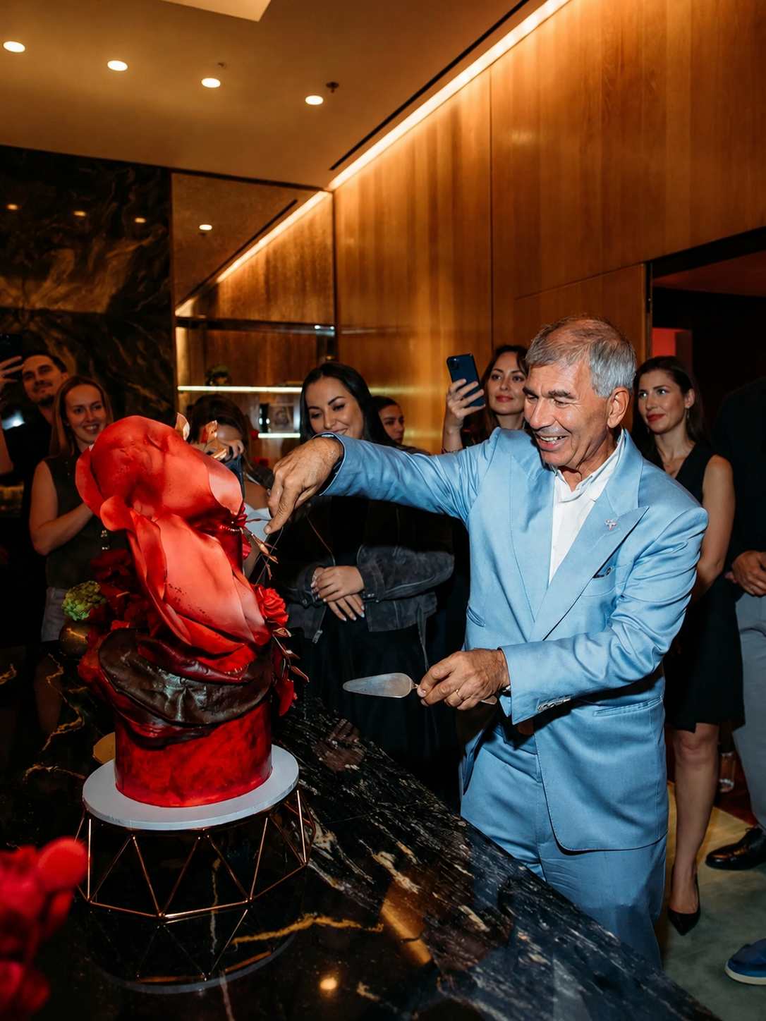 An elderly man in a light blue suit cuts a cake resembling a human heart during a celebration, surrounded by smiling guests taking photos.