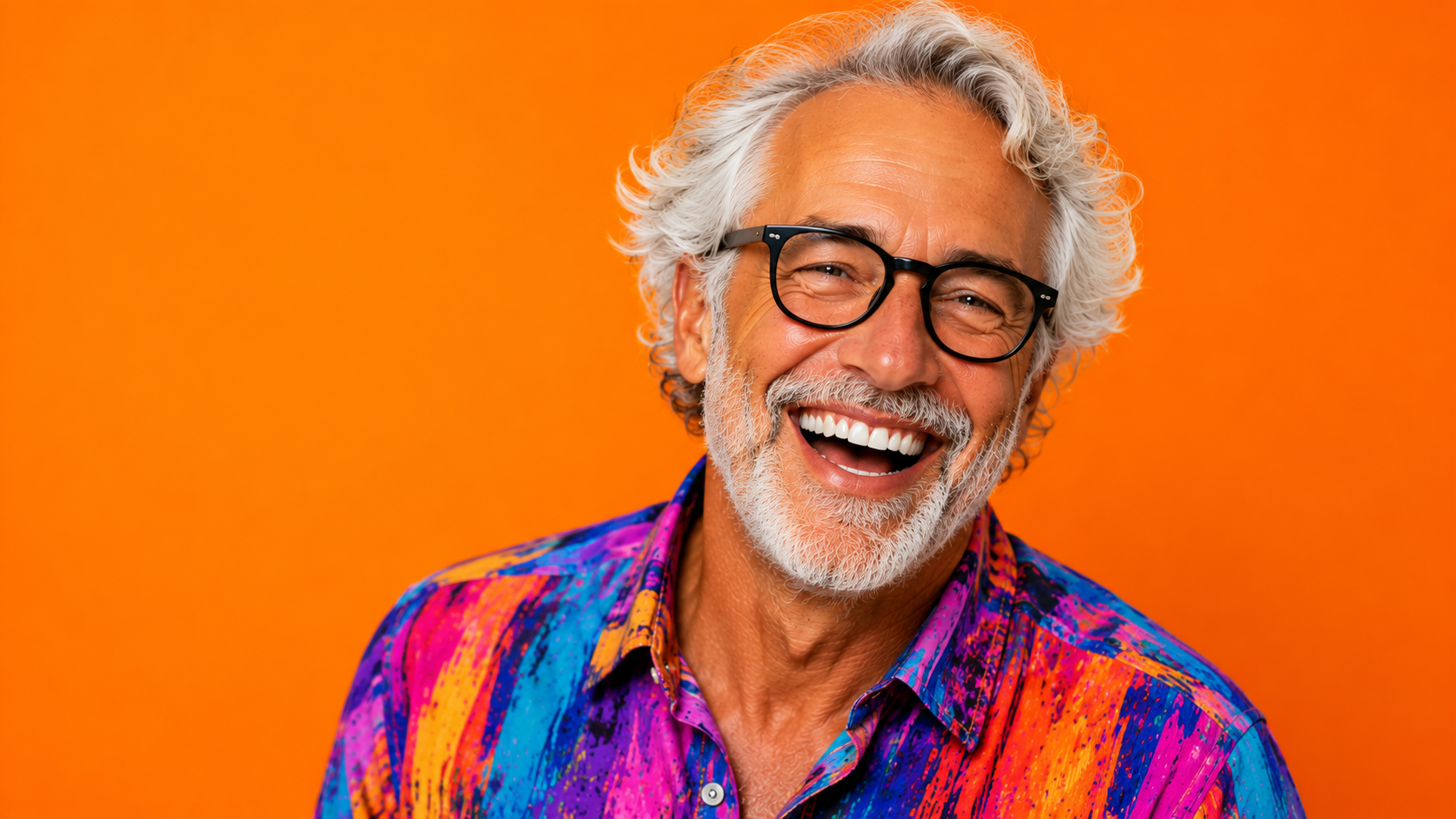 A cheerful smiling elderly man with gray hair, wearing black glasses and a colorful abstract-patterned shirt, standing against a vibrant orange background.