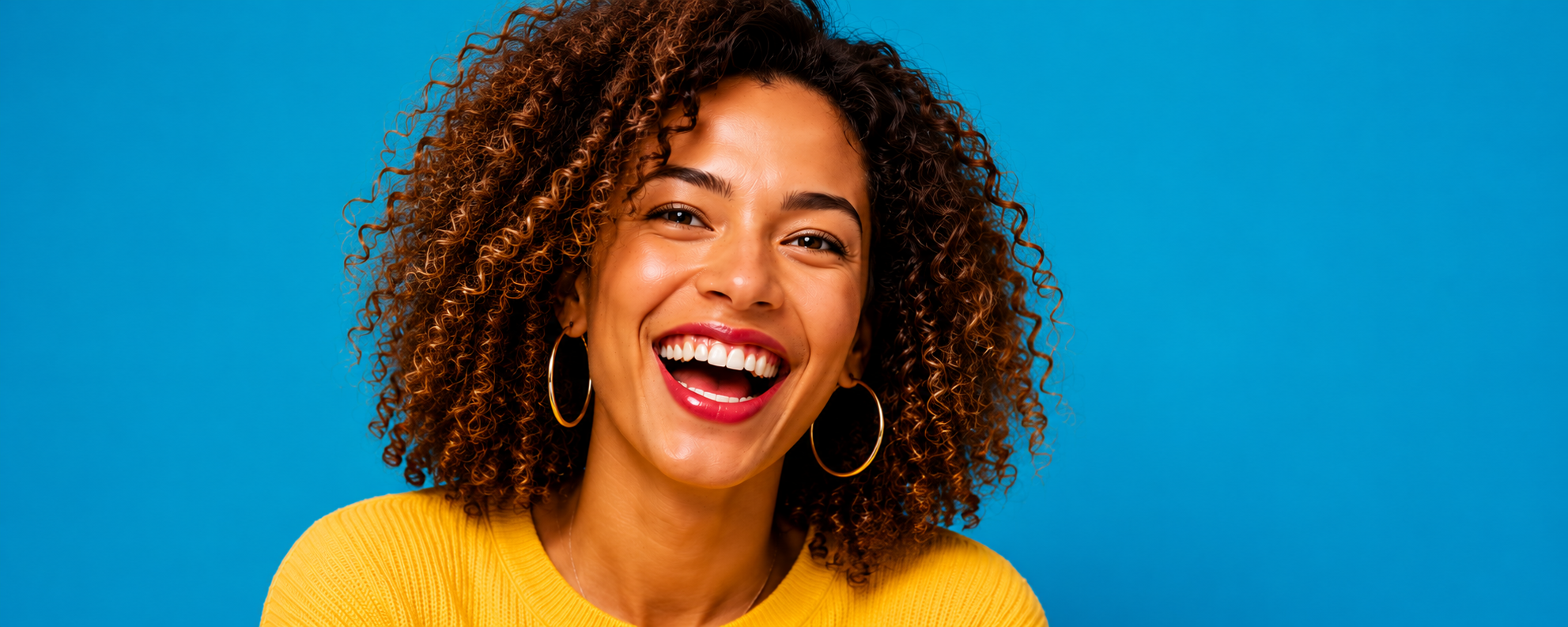 Close-up of a happy young woman with curly hair, wearing hoop earrings and a yellow top, smiling against a blue background.