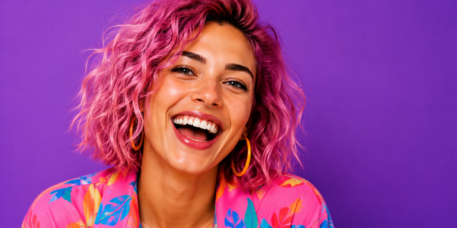 A woman with pink curly hair smiling widely, wearing yellow hoop earrings and a colorful floral shirt against a purple background.