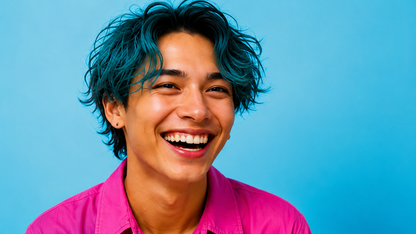 A person with short wavy hair dyed blue at the tips, smiling broadly against a light blue background, wearing a bright pink shirt.