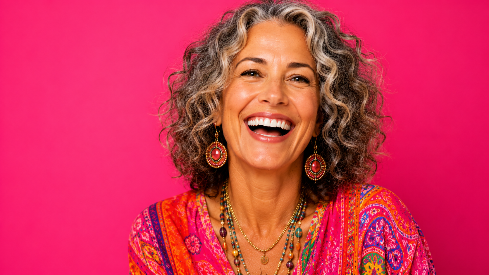 A smiling middle-aged woman with curly gray hair, wearing colorful bohemian jewelry and a vibrant pink patterned blouse against a solid pink background.