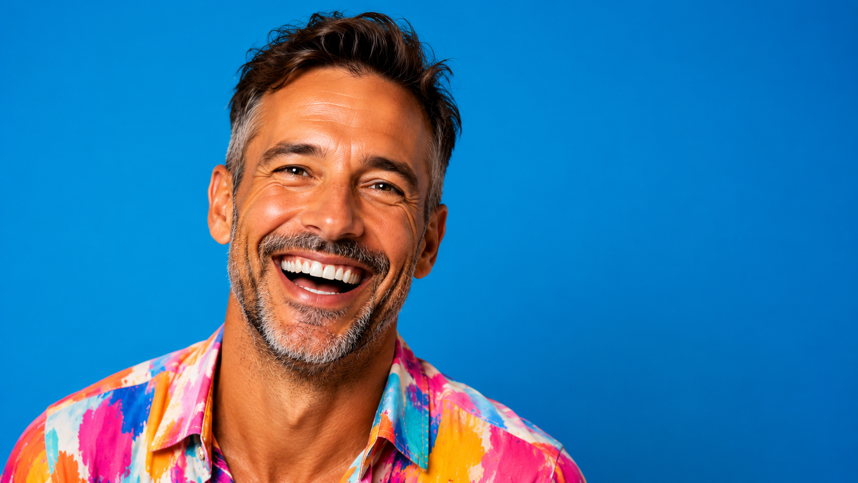 A middle-aged man smiling, wearing a colorful, paint-splattered shirt, with a blue background.