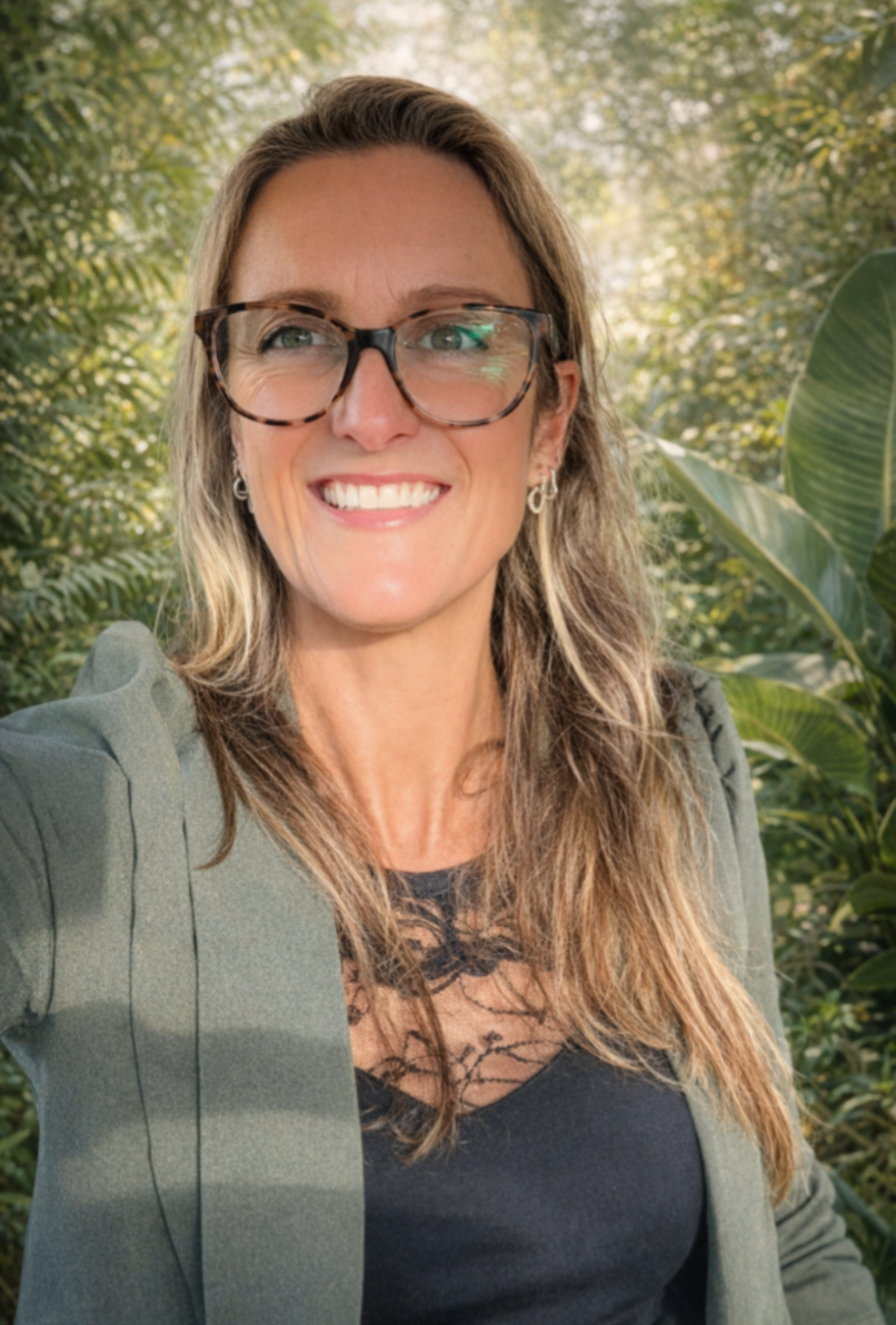 A woman with long, wavy hair and glasses taking a selfie outdoors with lush green foliage in the background.