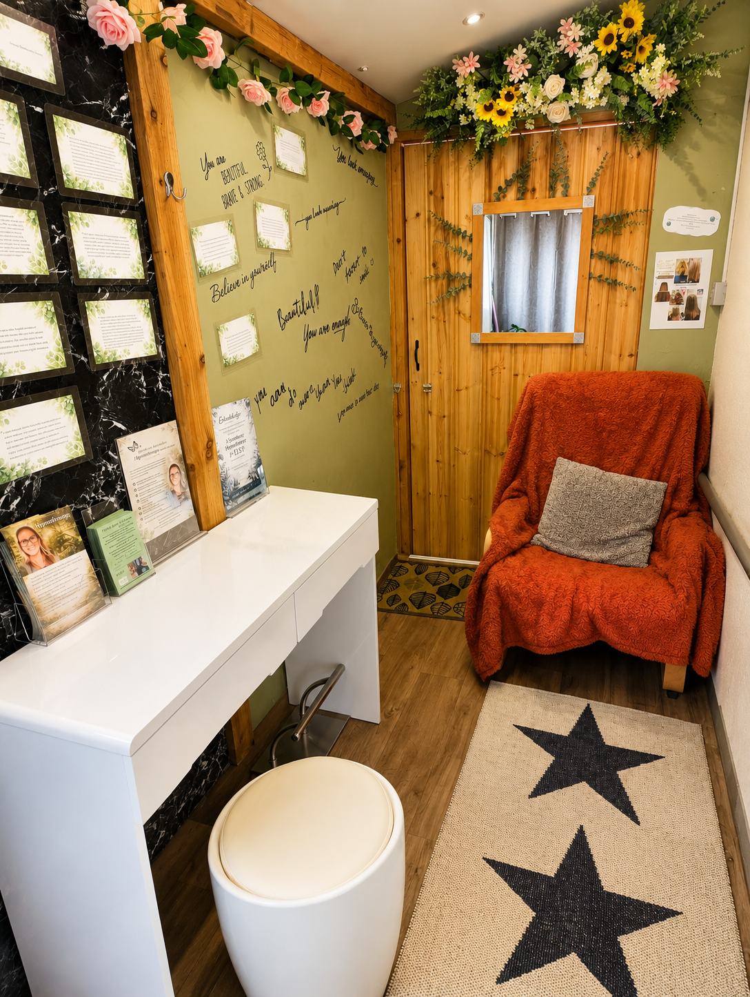 A cozy room with a green wall decorated with pink paper roses and inspirational quotes, a black marble display with informational cards, a white desk with brochures, a plush orange armchair with a gray pillow, a small mirror framed with greenery, a star-patterned rug, and a wooden door with yellow and white flowers above.