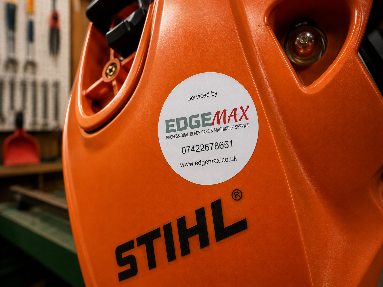 Close-up of an orange STIHL chainsaw with a white circular sticker that reads 'Serviced by EDGEMAX, professional blade care and machinery service, phone number, and website, on a workshop background.