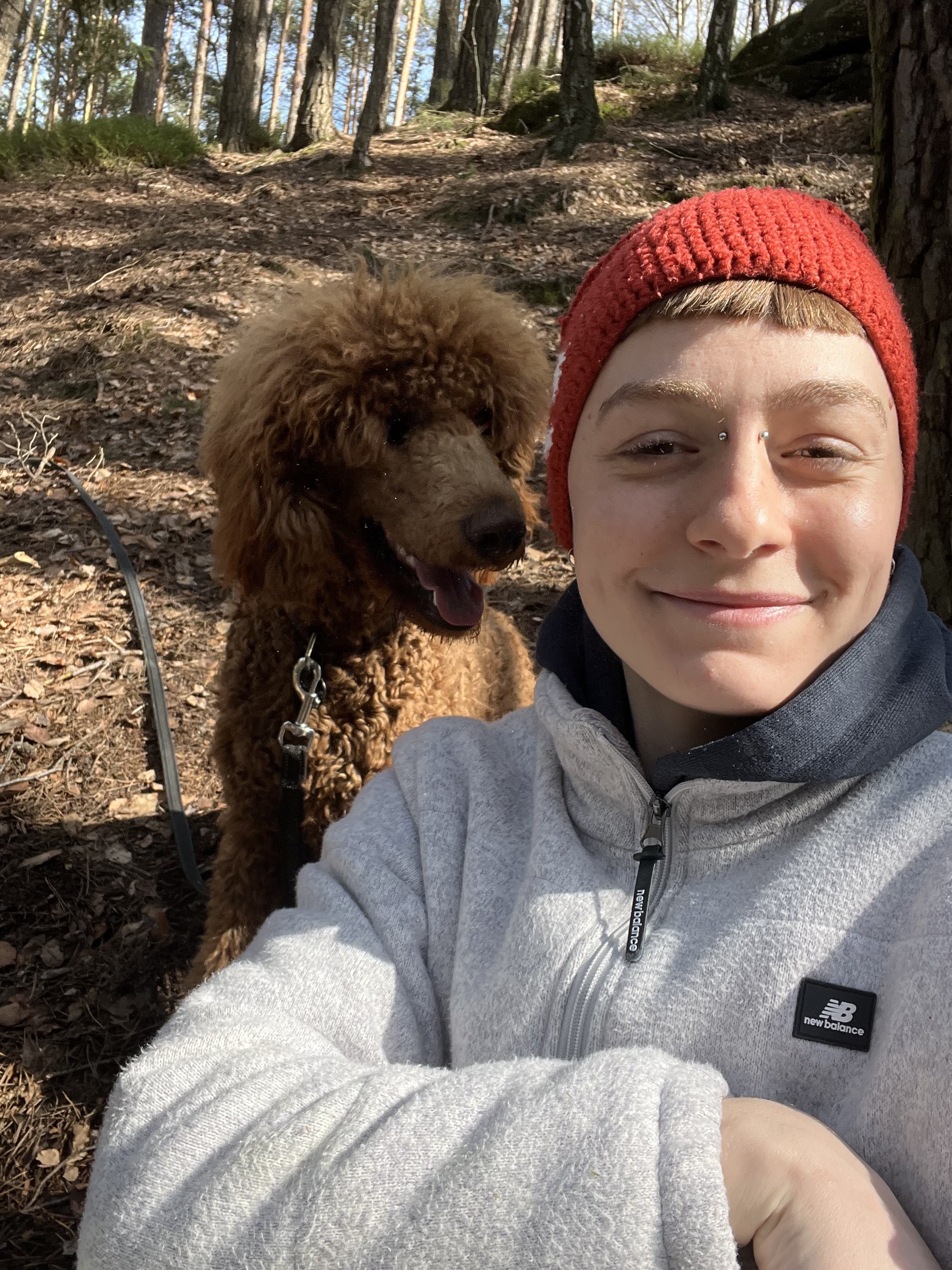 Ein Lächelnder Mensch mit Piercings im Gesicht fotografiert sich im Wald, neben einem braunen, lockigen Hund.