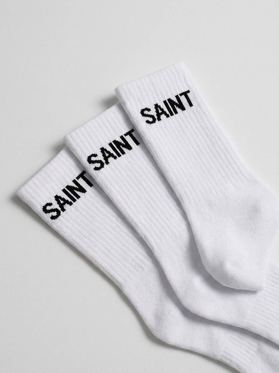 Two white athletic socks with black text that reads 'SAINT' on the ribbed top, laid on a white surface.