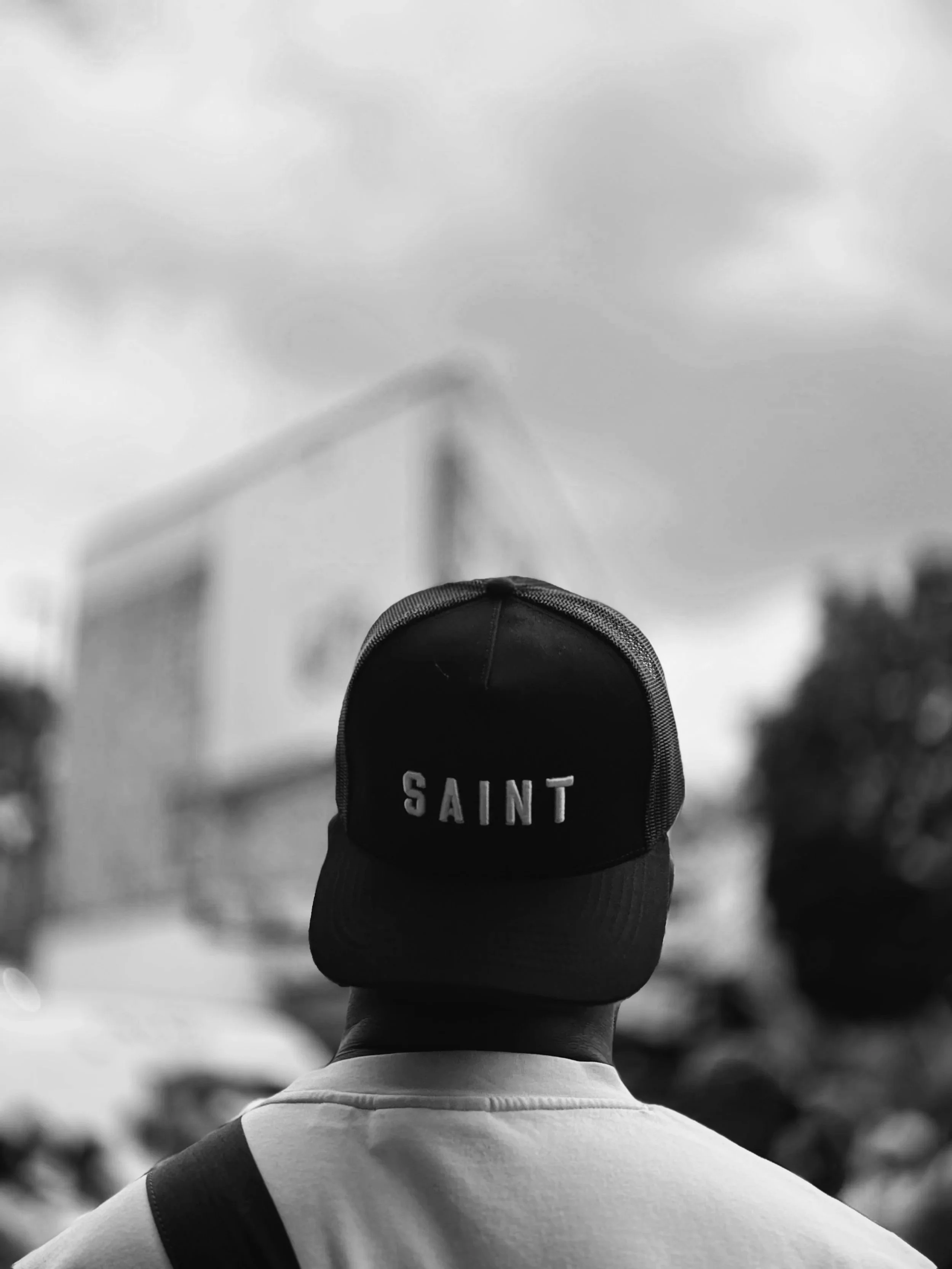 Back view of a person wearing a baseball cap with the word 'SAINT' embroidered on it, standing outdoors during cloudy weather.