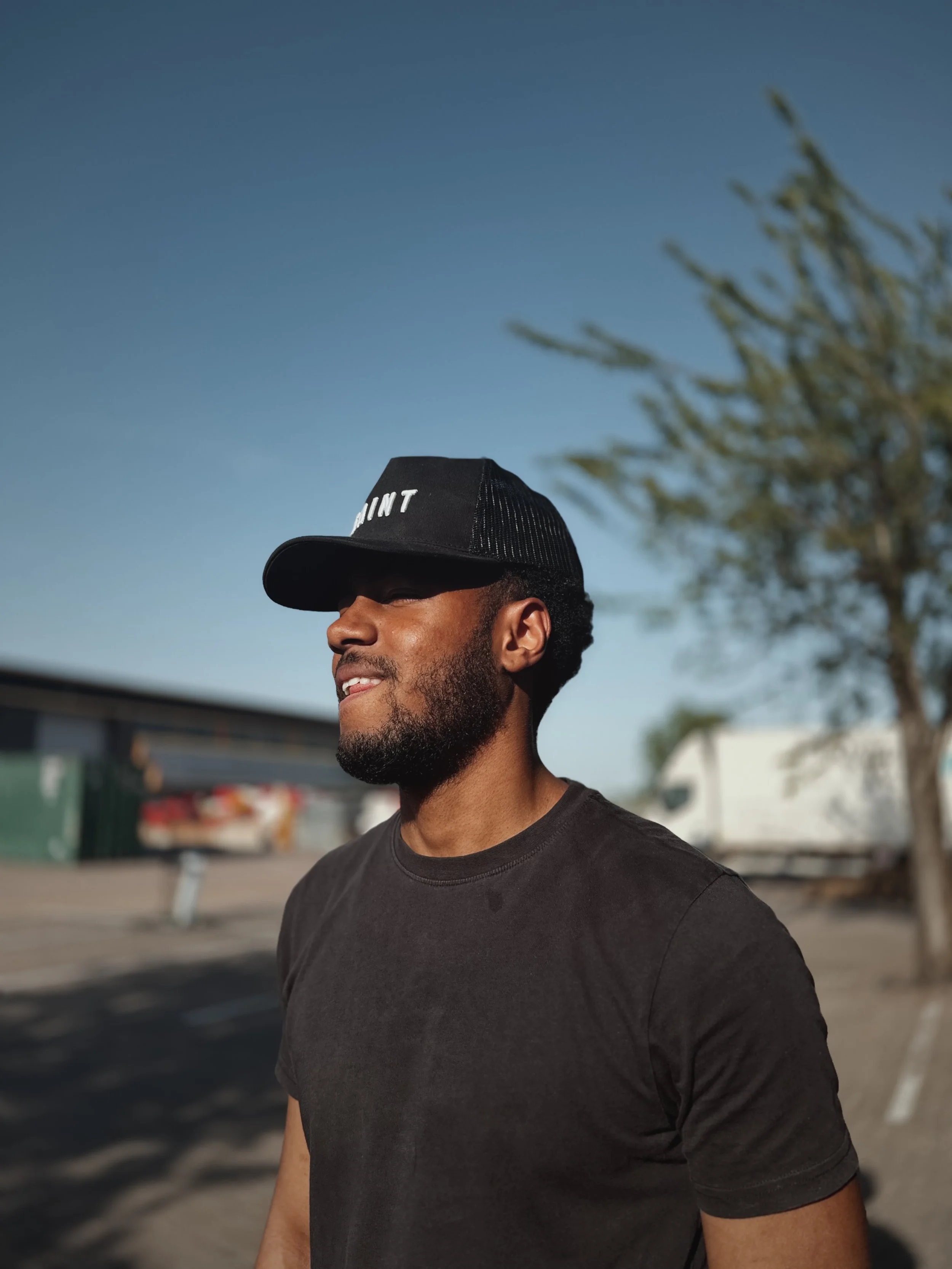 Keston with the Saint black trucker hat and a black t-shirt under a clear blue sky.