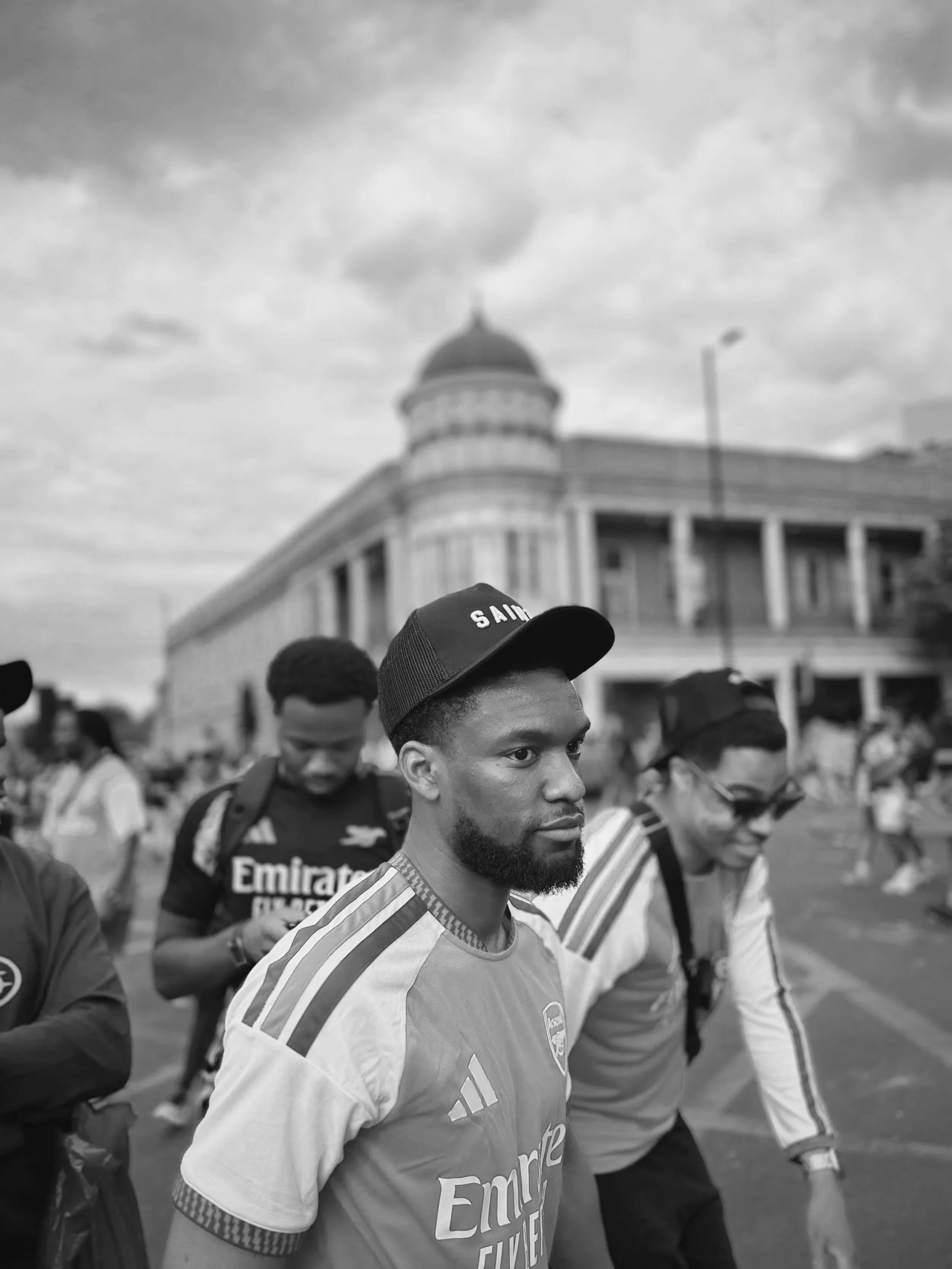 A black and white photo of a group of young men, with one in the foreground wearing a sports jersey and a baseball cap, walking in a line outdoors. In the background, there is a large building with a dome and sky with clouds.