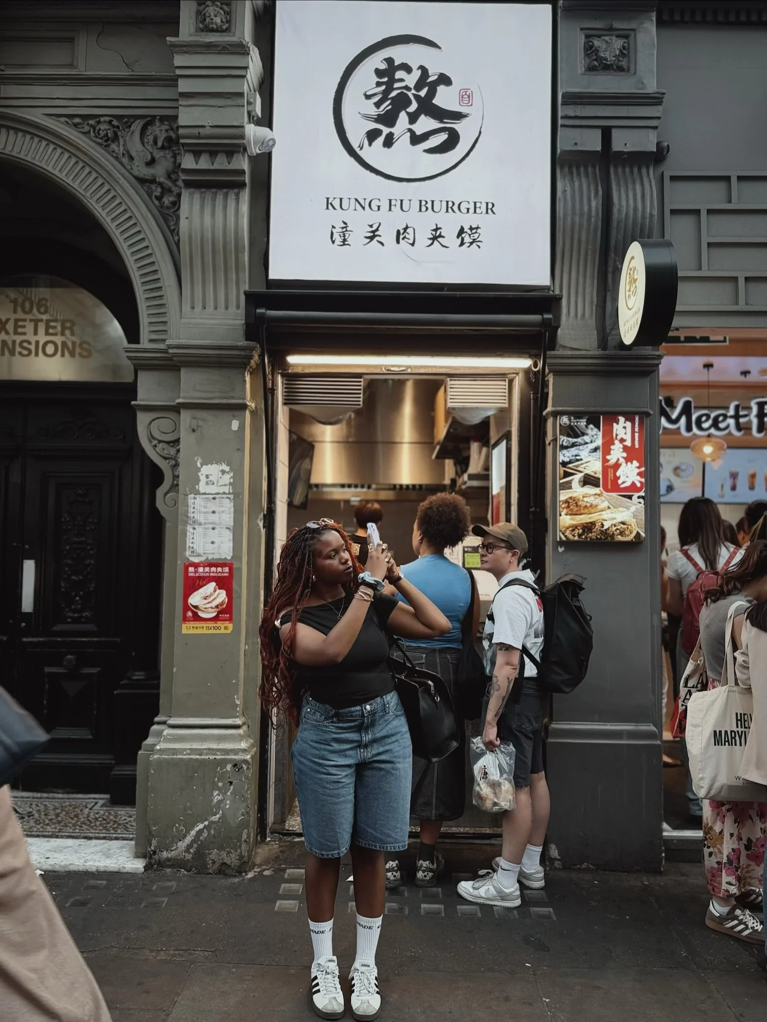 Seyi outside a restaurant called 'Kung Fu Burger' with Asian-inspired decor and signage, in front taking a photo or video with a smartphone.