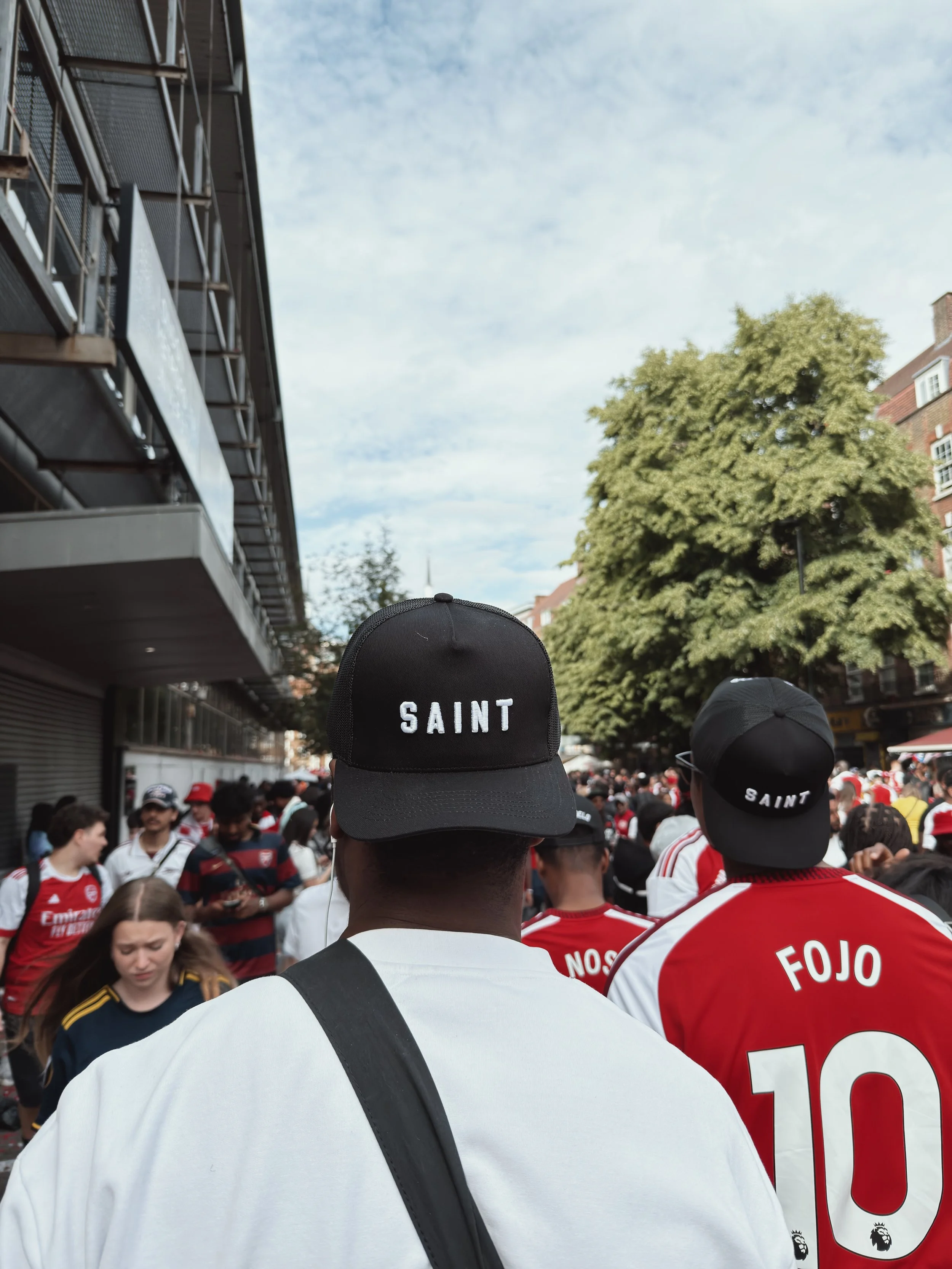 Crowd of people wearing football jerseys and caps, with two individuals in the foreground wearing black caps with "SAINT" written on them, in an outdoor city setting with trees and buildings.