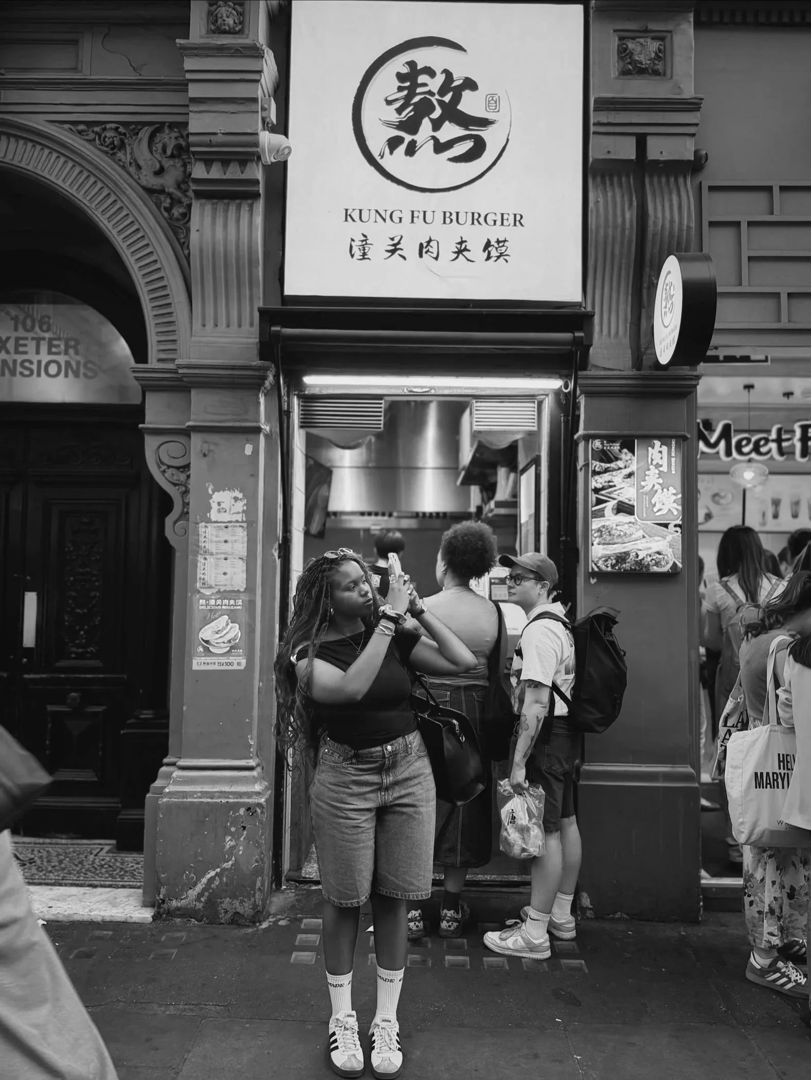 Black and white photo of people in front of a restaurant with a sign that reads "Kung Fu Burger" in English and Chinese characters. A woman with long braids is standing in the foreground, holding her phone and taking a photo of people in front of her.