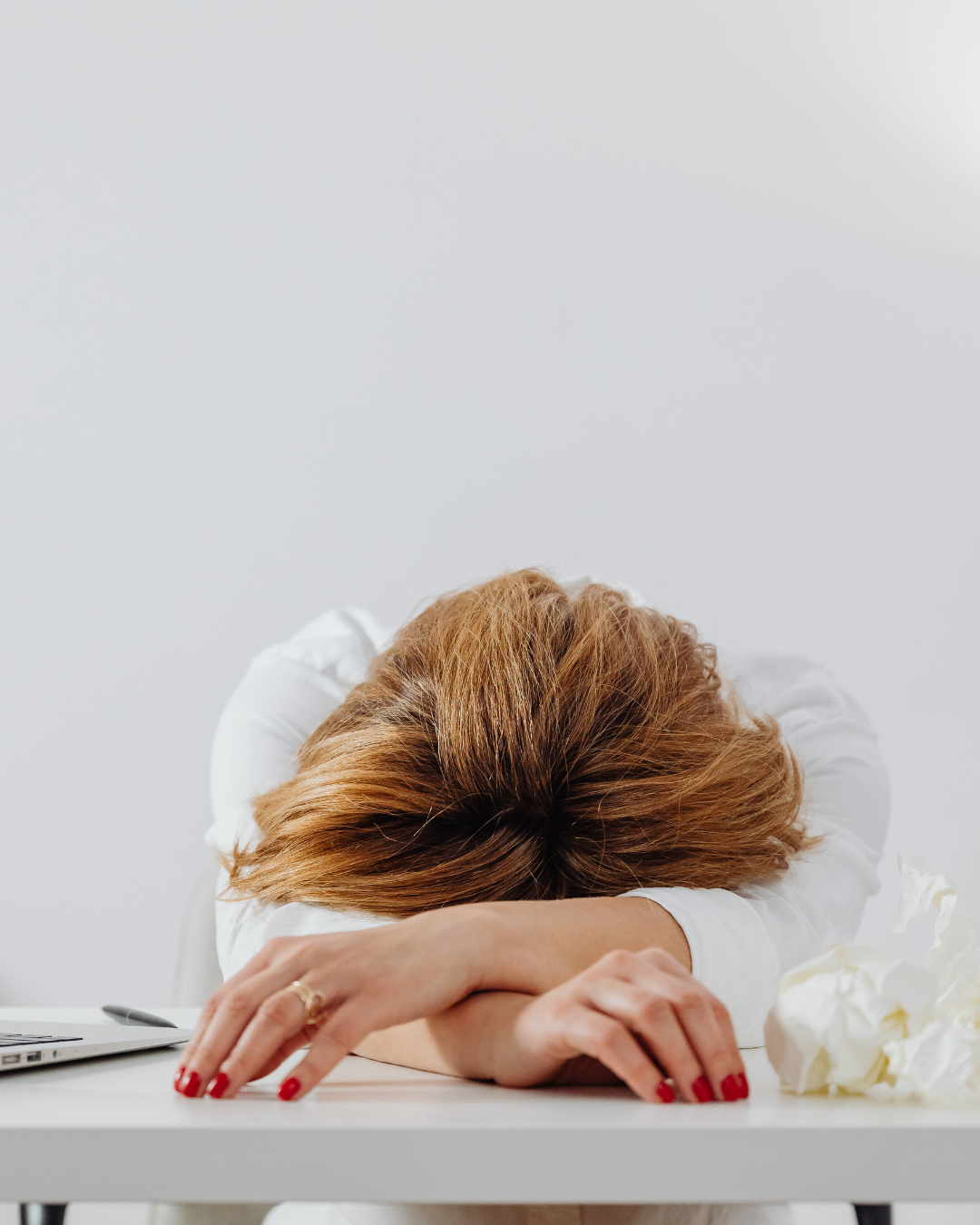 A woman with red hair resting her head on her crossed arms on a white desk, appearing exhausted or frustrated.