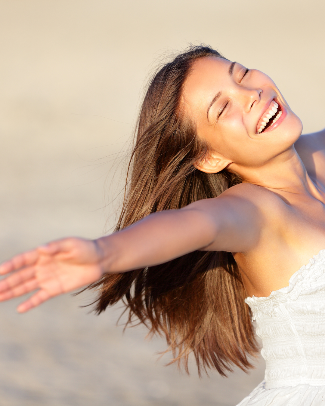 A woman with long brown hair smiling joyfully with her arms outstretched, outdoors, during sunset.