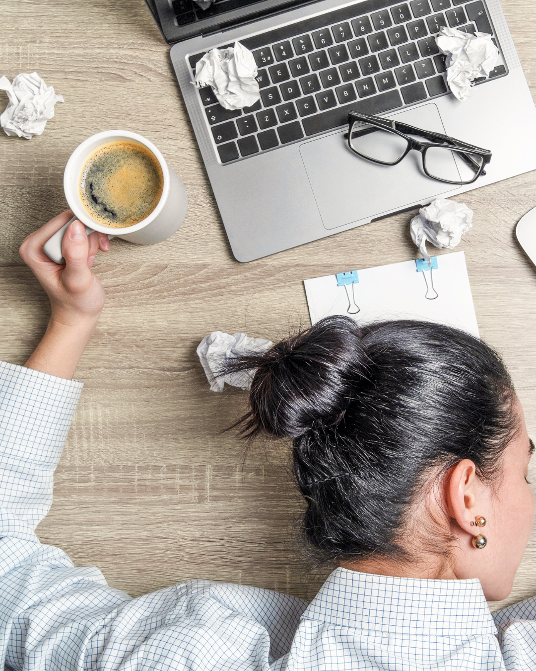 Cluttered workspace with a woman resting her head on a desk, surrounded by crumpled paper, a cup of coffee, a laptop with glasses on top, and a piece of paper clipped with two blue clips.