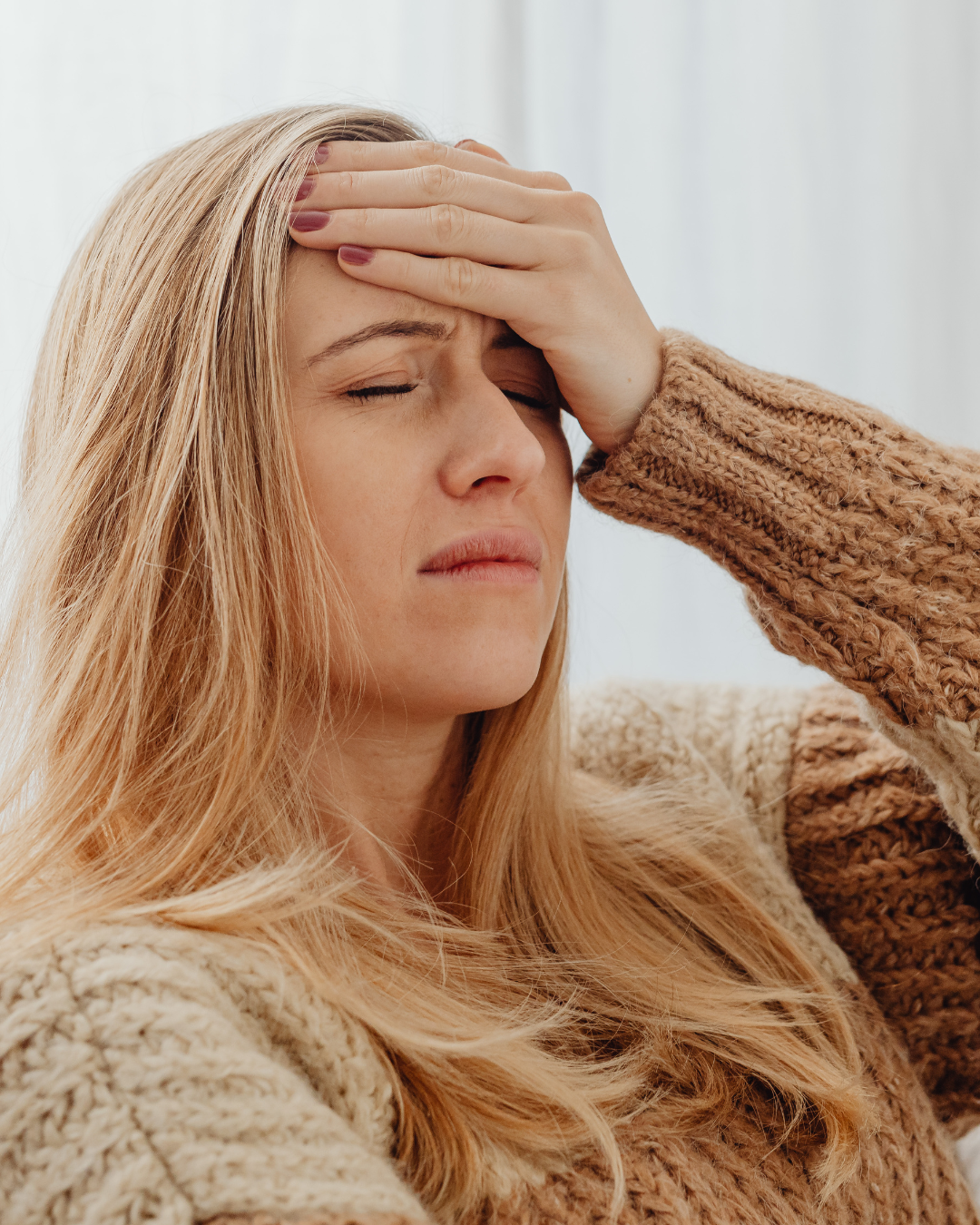 A woman with long blonde hair, wearing a beige knitted sweater, is holding her forehead with her hand and appears to be in pain or distress.