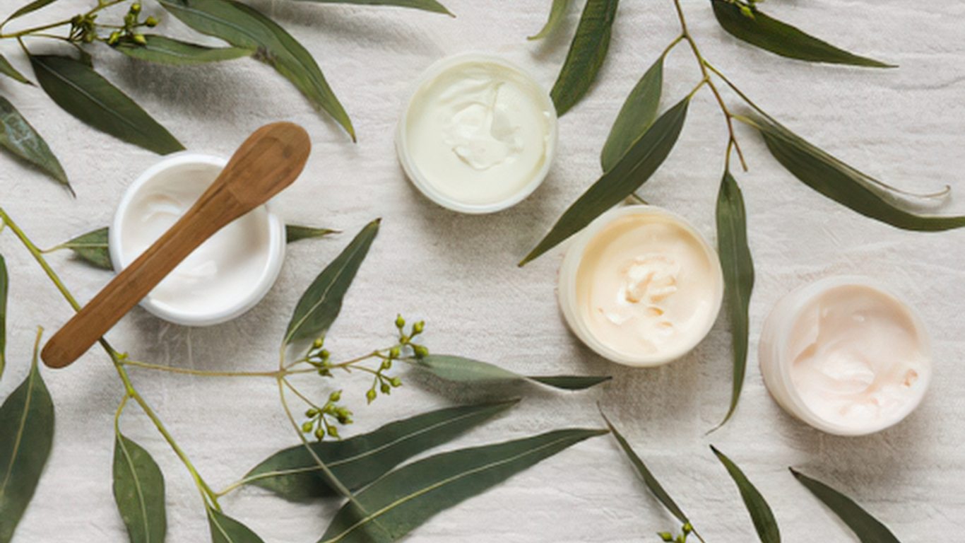 Four small open jars of cream or moisturizer surrounded by eucalyptus leaves and small green buds, with a wooden spatula resting on one of the jars, on a white textured surface.