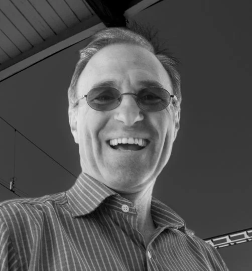 A man smiling, wearing sunglasses and a striped shirt, taking a selfie outdoors.