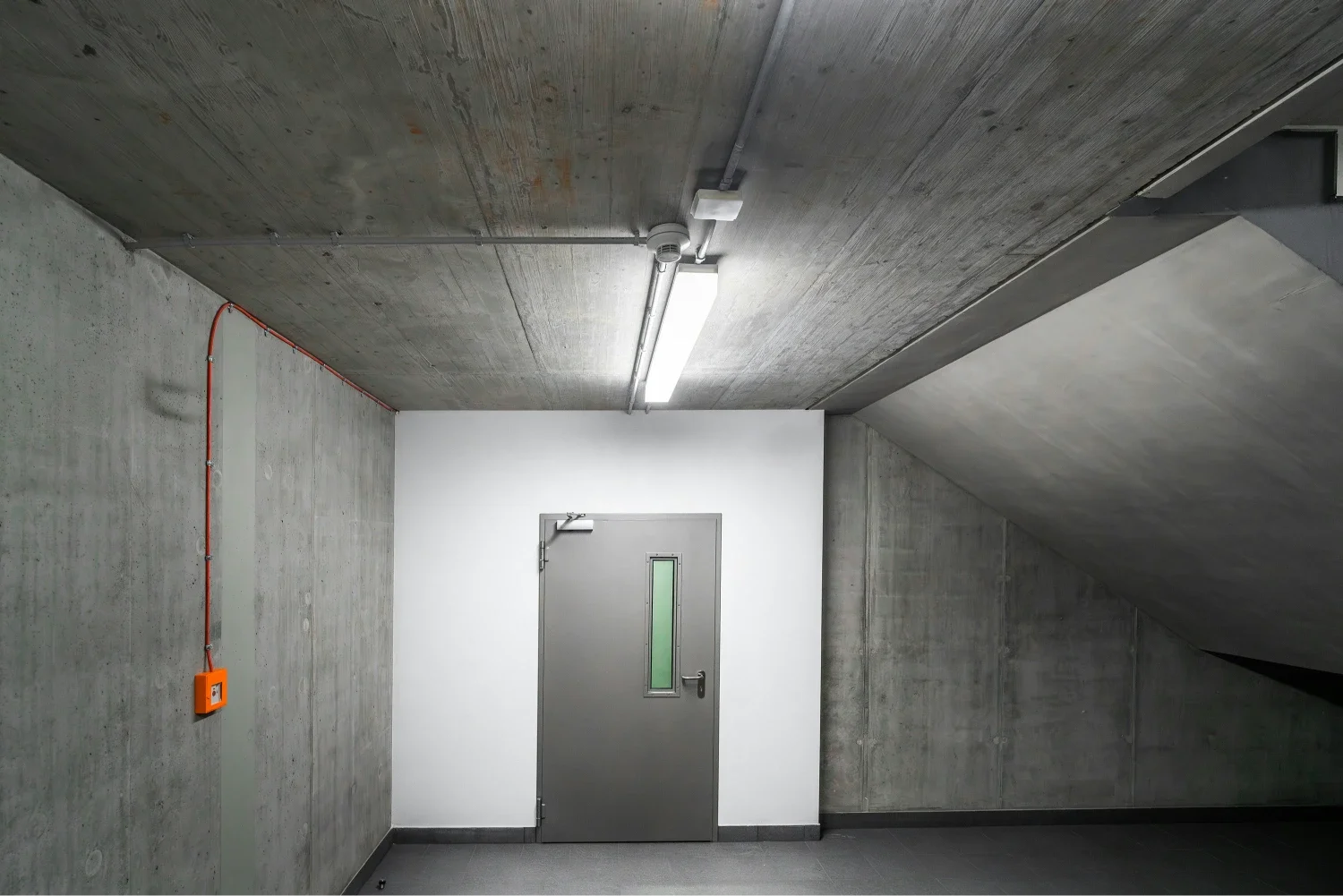 A plain, industrial-style hallway with concrete walls and ceiling, a gray metal door with a small window, a bright fluorescent light, and an orange electrical box mounted on the wall.