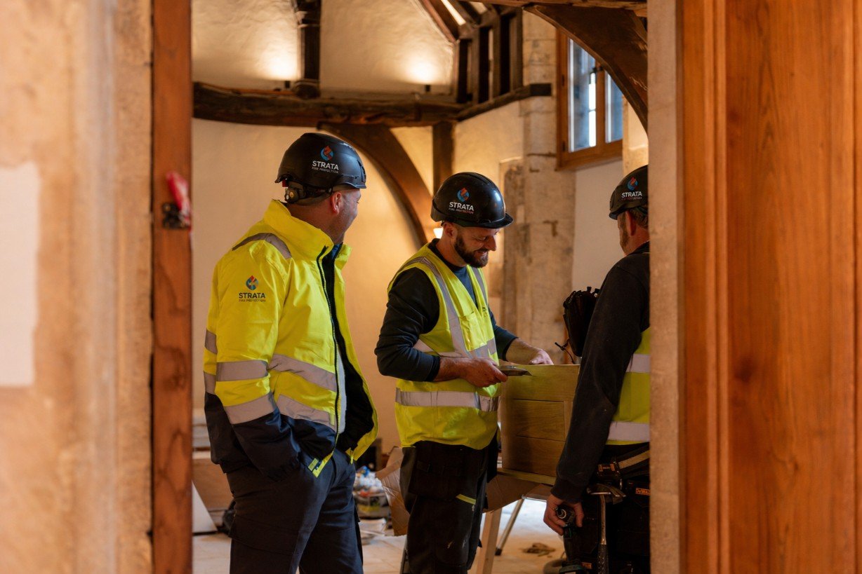 Three construction workers in yellow safety jackets and black helmets inside a rustic building with exposed wooden beams, engaged in a discussion over a clipboard.